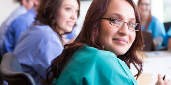 LatinX female nurse sitting at table smiling with colleagues staring at camera