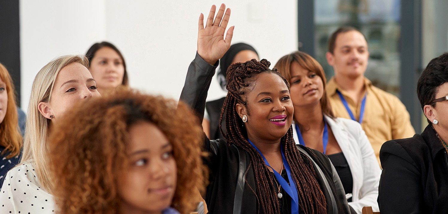 group of diverse people sitting at a conference with one woman's hand raised smiling, for a question